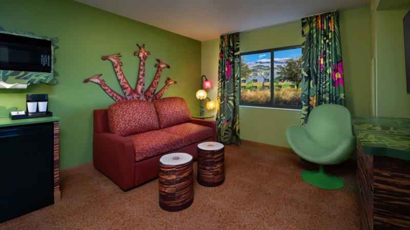 Interior of a room with laminate flooring, an upholstered chair, 2 ottomans, a couch, a wall mounted flat screen TV, a side table, a dresser, a microwave, a mini fridge, curtains and a window
